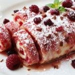 Eine hellrosa Himbeer-Joghurt-Rolle, die elegant auf einem weißen Teller präsentiert wird. Die Rolle ist fein geschnitten, zeigt eine cremige Füllung mit frischen Himbeeren, und die Oberfläche ist smooth mit einer leichten Puderzuckerüberziehung.