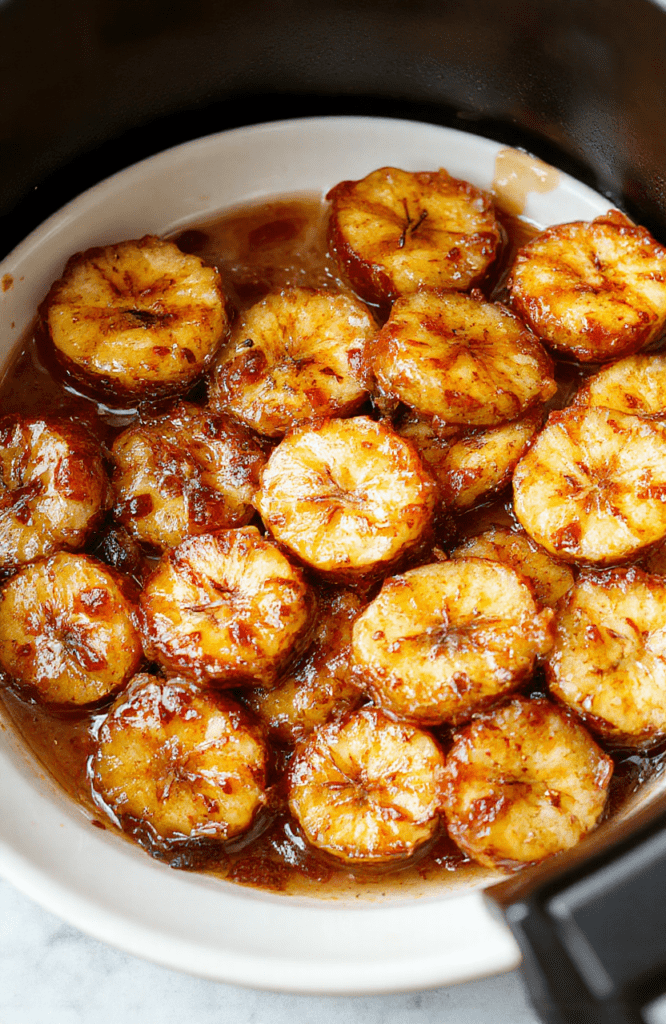 A close-up shot of perfectly caramelized bananas sliced and arranged on a white plate with a golden, glossy caramel coating. The bananas are topped with a sprinkle of cinnamon and drizzled with additional caramel sauce. The background features a softly blurred kitchen setting with warm lighting, emphasizing the inviting texture and rich color contrast of the dessert.