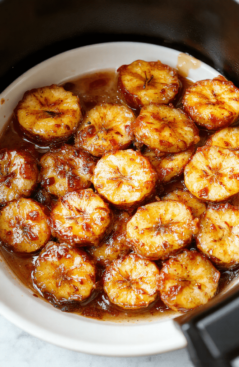 A close-up shot of perfectly caramelized bananas sliced and arranged on a white plate with a golden, glossy caramel coating. The bananas are topped with a sprinkle of cinnamon and drizzled with additional caramel sauce. The background features a softly blurred kitchen setting with warm lighting, emphasizing the inviting texture and rich color contrast of the dessert.